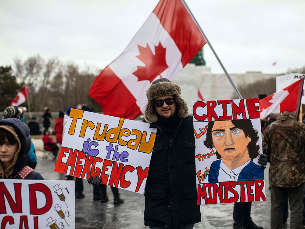 Protesters stand outside on a grey day. A man in a fur hat holds a sign that says 'Trudeau is the emergency' and 'Crime Minister.'