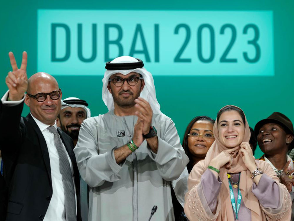 From left: a bald man with medium skin tone, glasses and a dark suit and grey tie holds up his hand making a V sign; a man with medium skin tone, glasses and a white keffiyeh applauds; a woman with medium-light skin tone and a beige hijab smiles. Behind them are the words 'DUBAI 2023' on a green screen.
