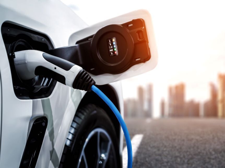 The charging port of a white EV with a charger plugged in. A blurry, sunny cityscape can be seen in the background.