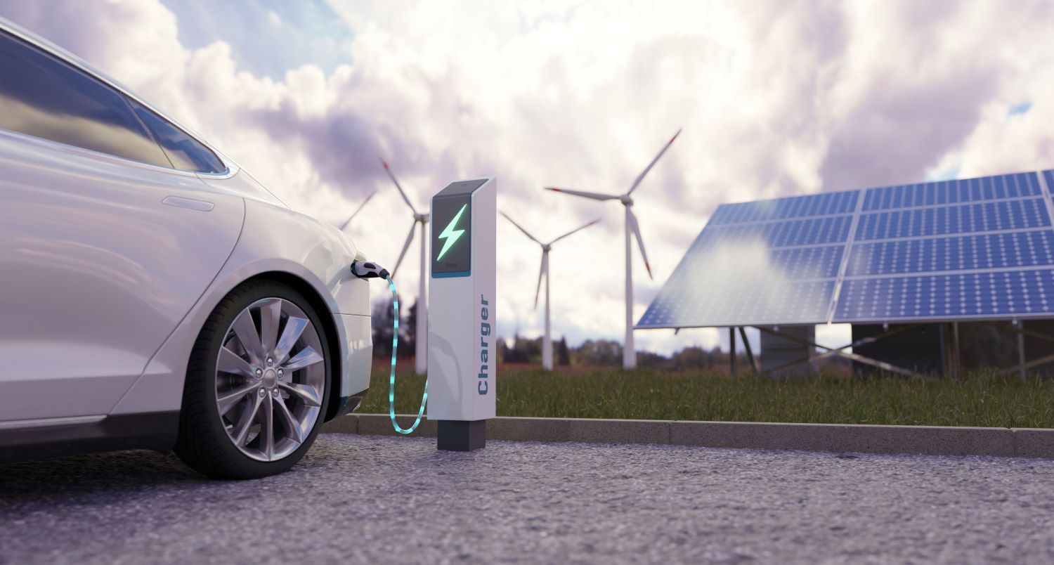 A white EV is plugged into a charging station with a lit up lightning bolt and the word 'Charger' on it. In the background are wind turbines and a large solar panel.