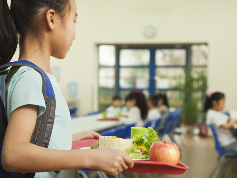 A student wearing a blue backpack is holding a food tray that includes salad and an apple in a school cafeteria.