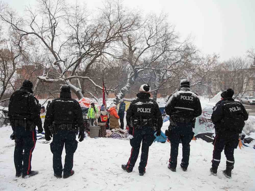 Five police officers in black uniforms stand with their backs to the camera on a snowy day. In front of them people stand, with tents and snow-covered leafless trees in the background.