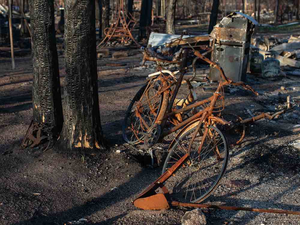 A burned bicycle sits on scorched ground, beside blackened trees in front of destroyed household possessions.
