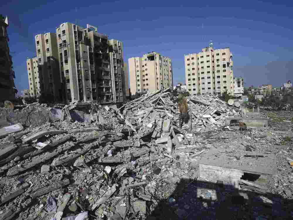 Piles of grey rubble fill the foreground. In the background are light beige highrise apartment buildings against a blue sky.