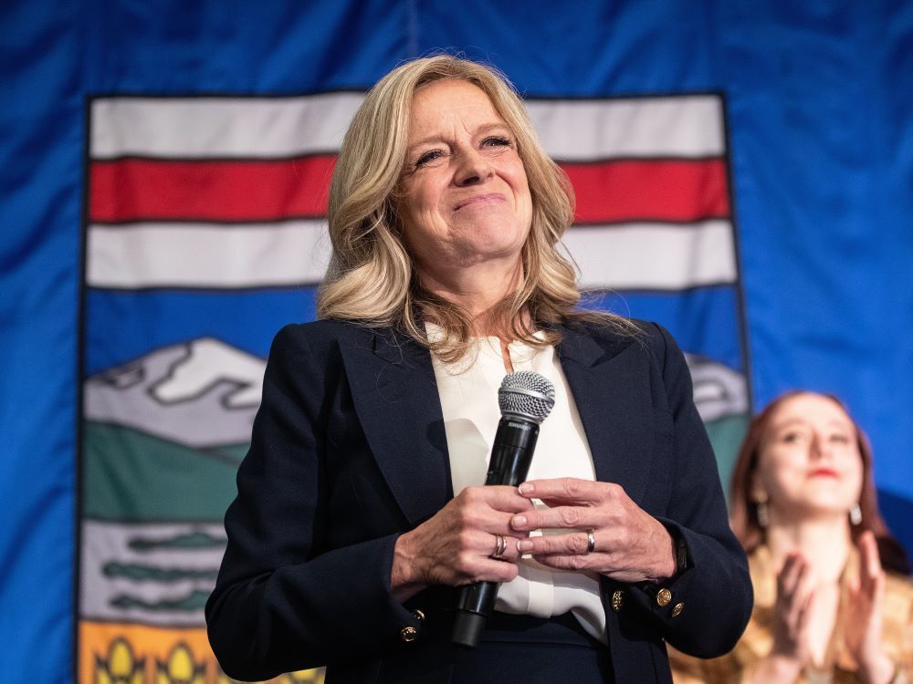 A blond white woman in a white shirt and dark jacket holds a microphone. She’s smiling and looks on as a woman behind her claps. The flag of Alberta hangs as her background.