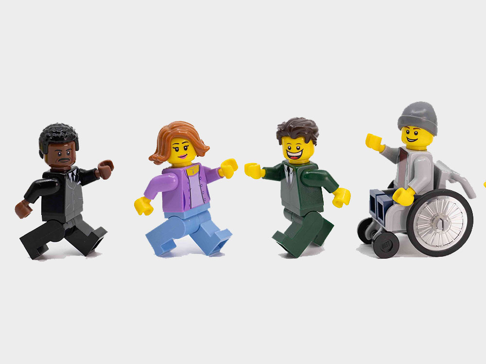 A row of Lego figurines stands across a white background. They represent a range of ages, abilities and ethnicities, and are posed to look like they are mid-step.