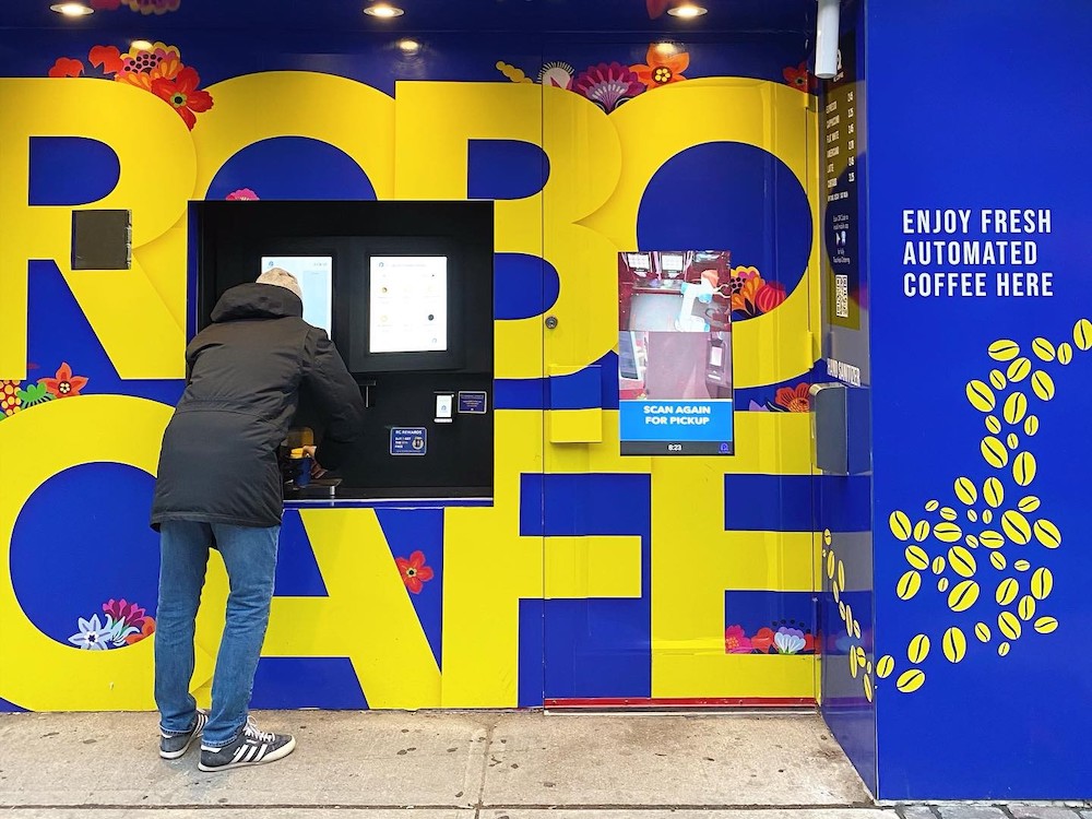 A person gets coffee from a street-side kiosk with a royal blue background and the words 'Robo Cafe' painted in yellow. To the right of the frame, white lettering reads 'Enjoy fresh automated coffee here.'