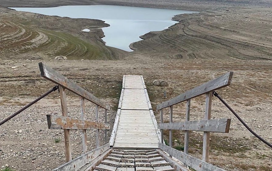 A wooden ramp juts out over a barren landscape of dirt with a small body of water in the distance.