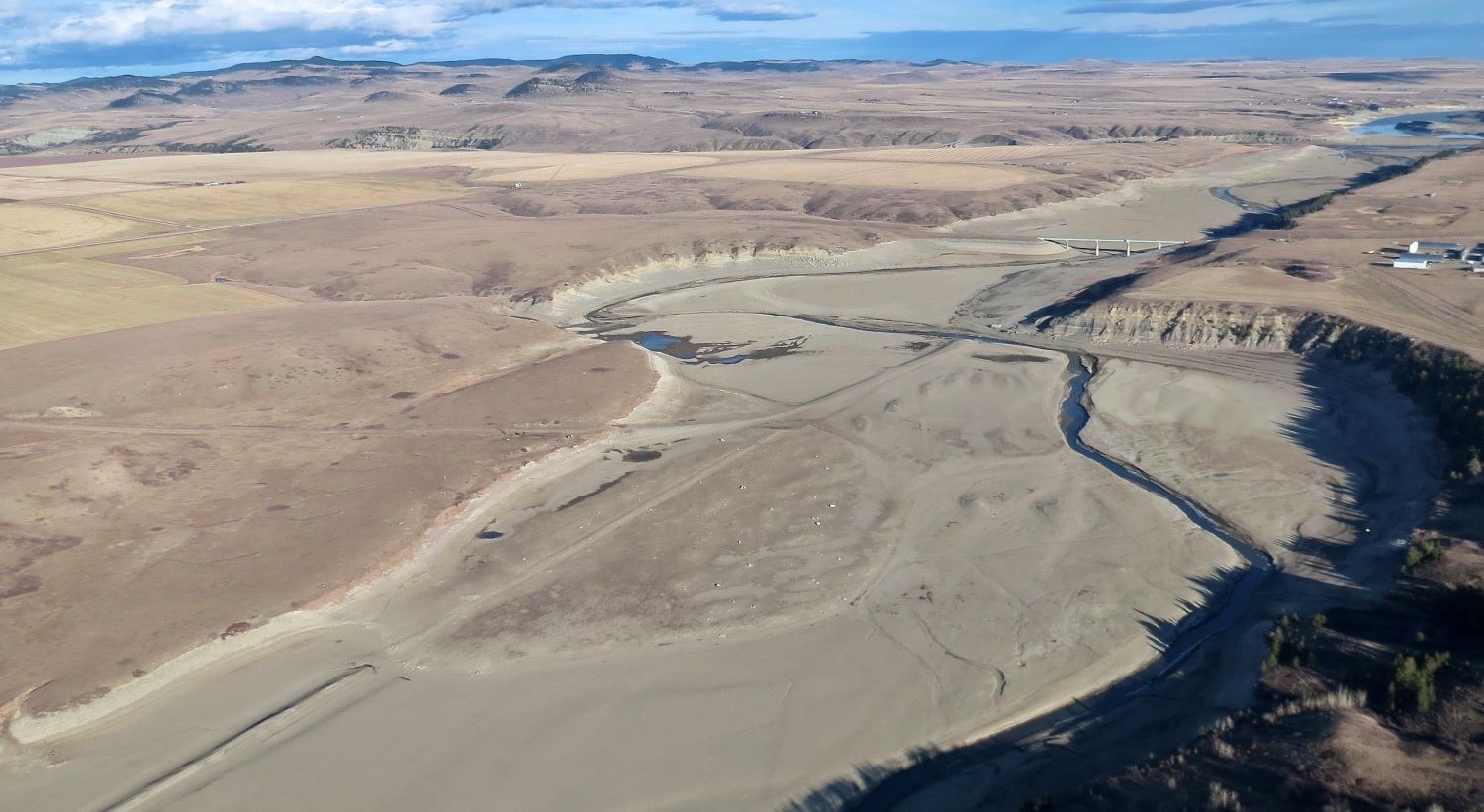 An aerial photo shows a tiny stream of water snaking through a vast landscape of barren brown soil. The previous outlines of a once wide river feeding into a reservoir are visible.