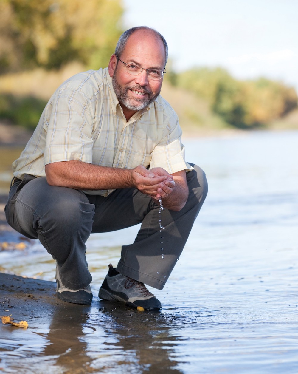 A light-skinned man with grey thinning hair, wearing an off-white shirt and grey pants, crouches on the bank of a river. Has palms are cupped together and water dribbles from them.