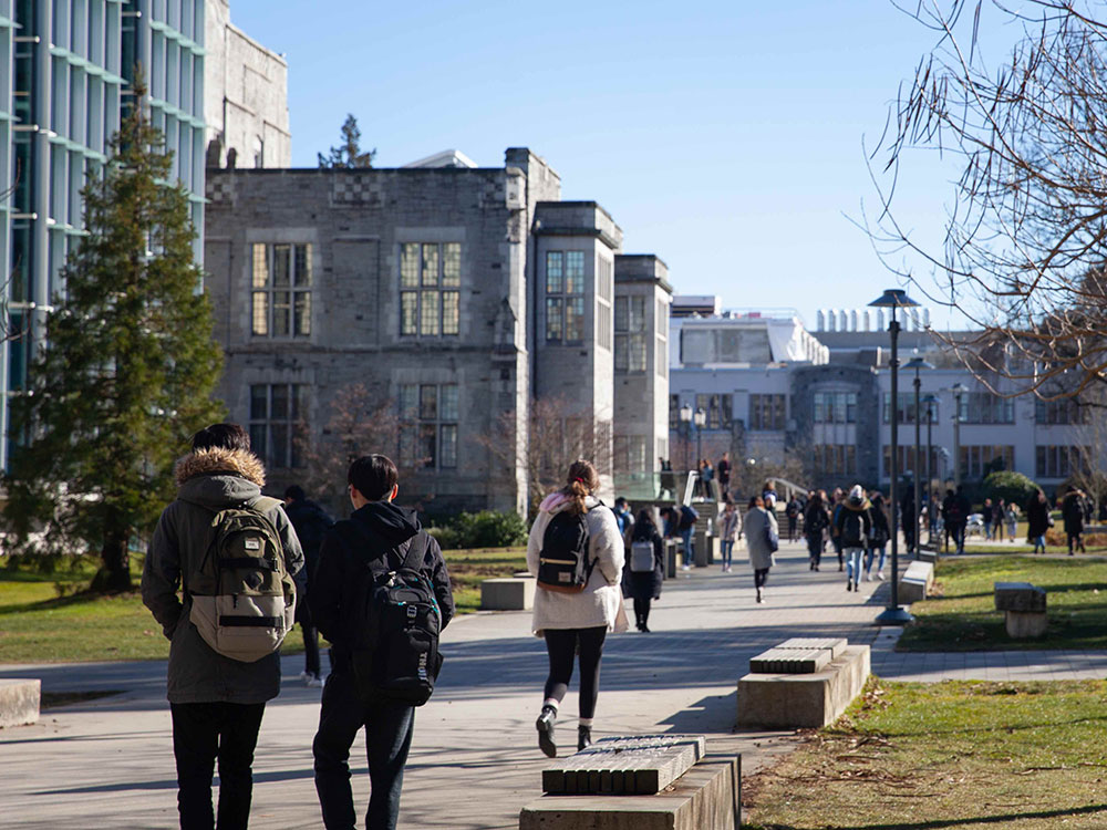 Young adults wearing winter jackets and backpacks walk along a concrete walkway beside a grey historic building at the University of British Columbia on a sunny winter day. Their backs are turned to the camera.
