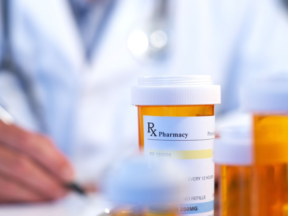 An RX prescription drug bottle is in focus while out of focus and in the background a doctor in a white coat writes a prescription.