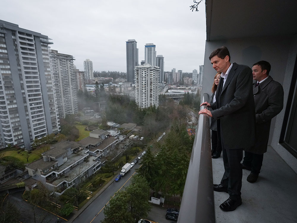 A very tall light-skinned man stands on a balcony looking down to the street and a green space. Two other people stand behind him. It’s a grey day.