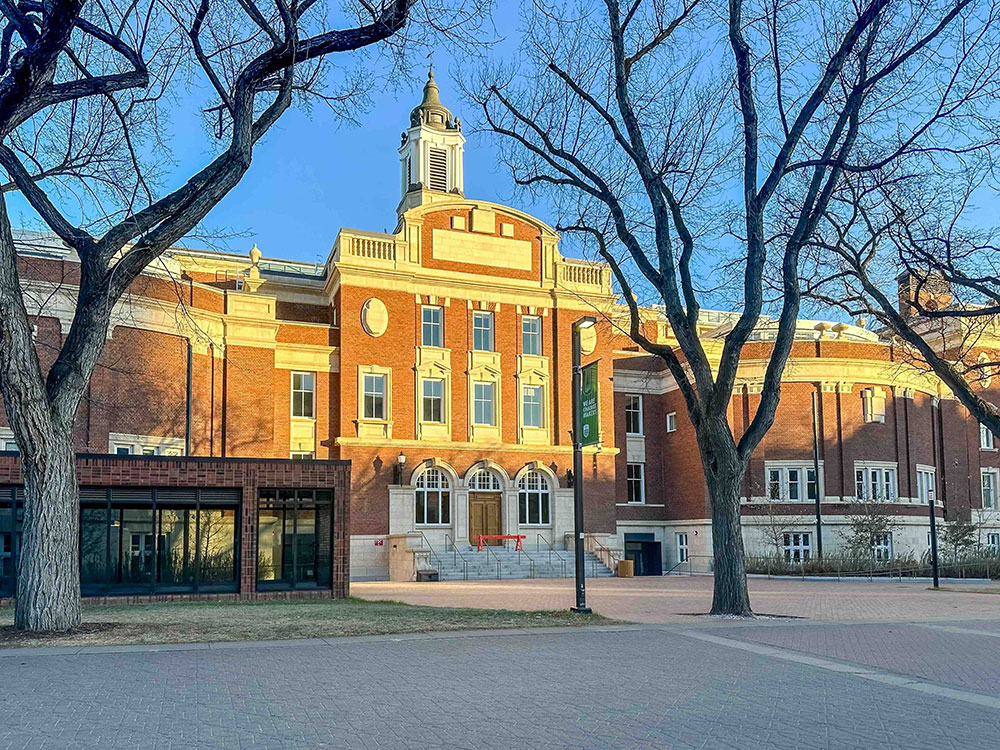 A historical building brown brick building with white trim on the University of Alberta campus at sunset. It is surrounded by bare winter trees and blue sky.