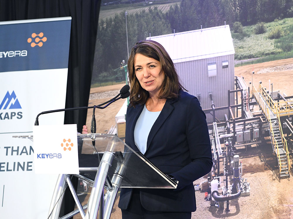 A 50-ish woman with shoulder length brown hair stands at a podium, wearing a dark blue suit and aqua shirt. Behind her is a photo of pipeline infrastucture.