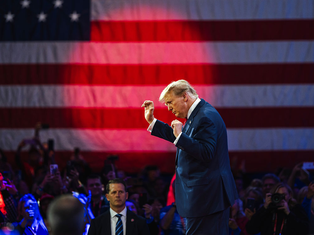 Trump dancing on a stage, with an American flag and a red spotlight. A crowd watches.