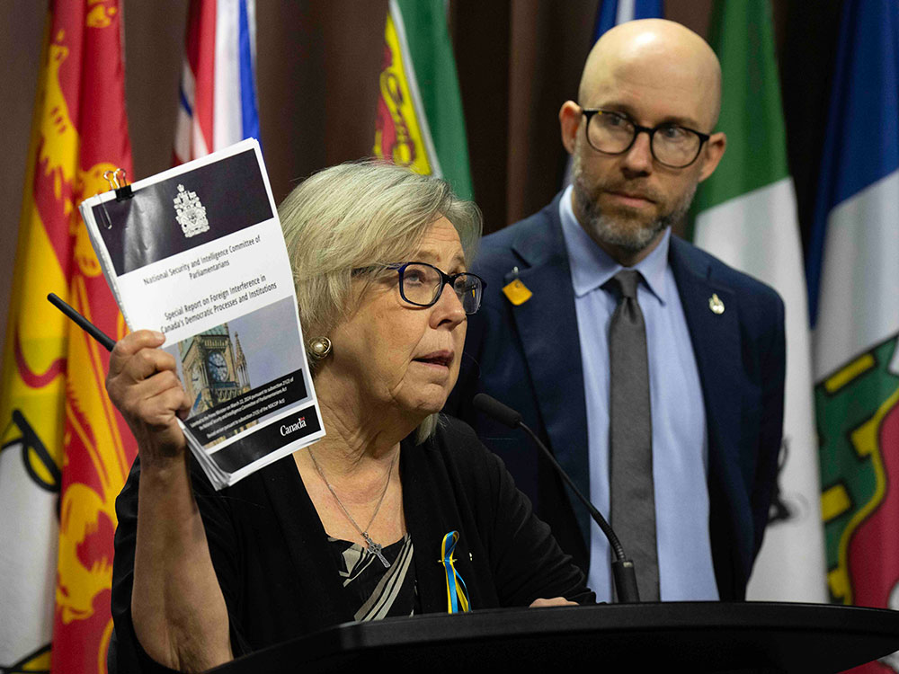 Elizabeth May, a woman in her early 70s, stands at a podium in front of the colourful flags of several Canadian provinces. She has light skin, blond hair, pearl earrings and glasses. She holds up a public security report while speaking. To the right of the screen in the background is Mike Morrice, a taller man in his late 30s. He has light skin, is bald with a beard and is wearing glasses with a blue suit.