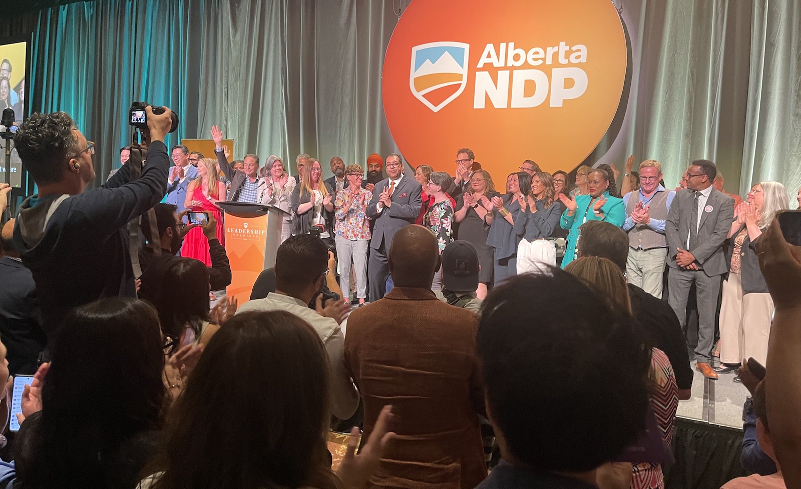 Before a clapping audience, a man with dark hair, dark-rimmed glasses and medium-toned complexion stands with dozens of people of different ages and ethnicities on a stage with an 'Alberta NDP' sign behind them.
