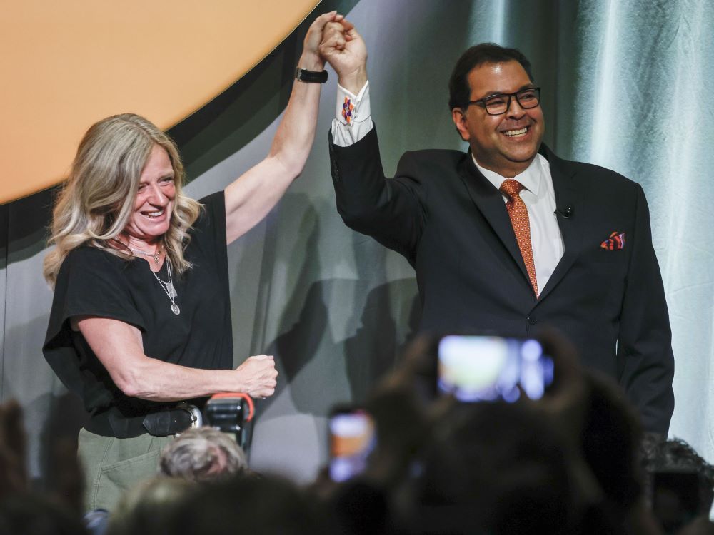 A blond-haired, middle-aged light-complexion woman raises joined hands with a suit and tie-wearing man with dark hair, dark glasses and medium complexion. Both are smiling.