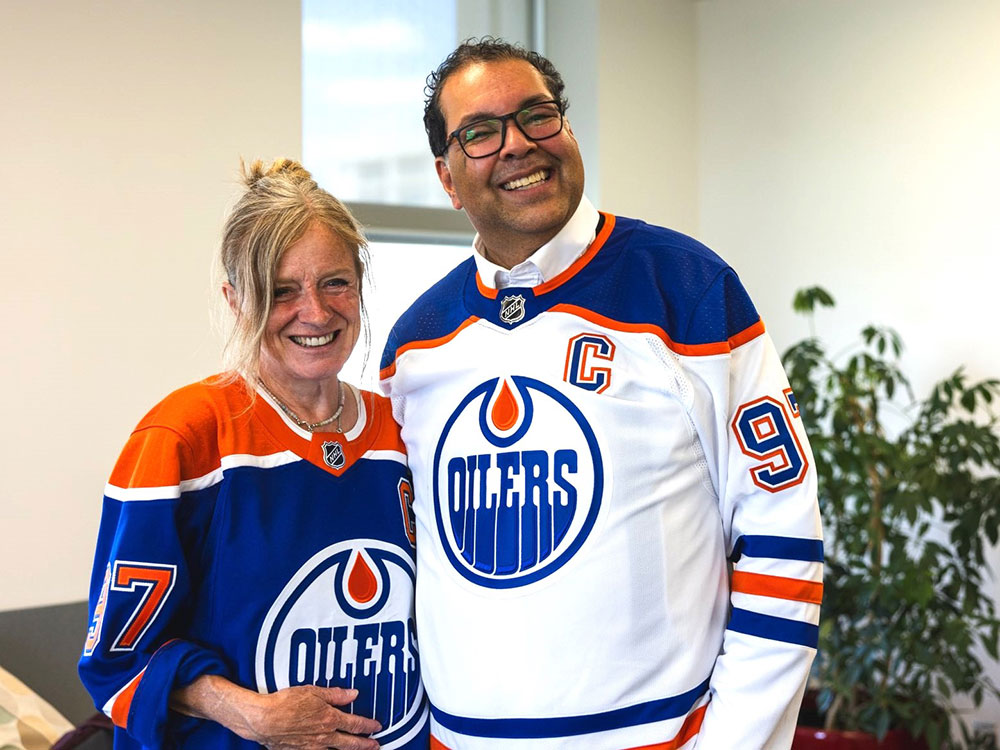 Naheed Nenshi and Rachel Notley wearing Edmonton Oilers jerseys. Notley, at left, is wearing the orange and blue home colours. Nenshi, at right, is wearing the white with blue accent away colours.