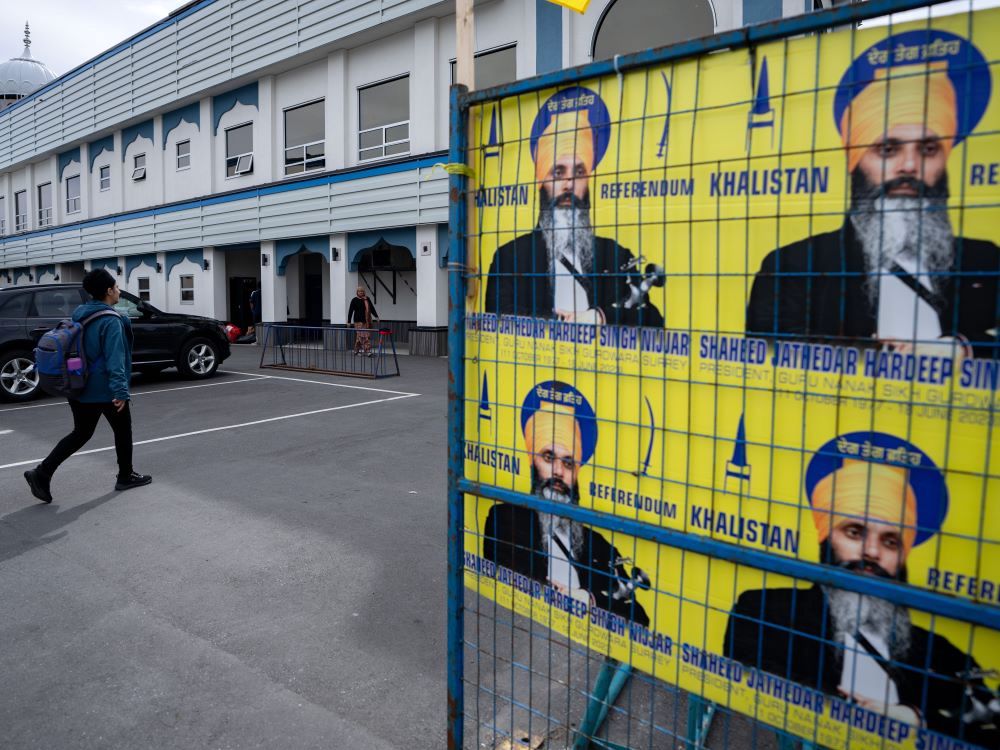 Outside a gurdwara, yellow signs attached to a blue metal frame show a man with medium skin tone, a black moustache and a long grey beard wearing a yellow turban, with the words 'Khalistan referendum.'