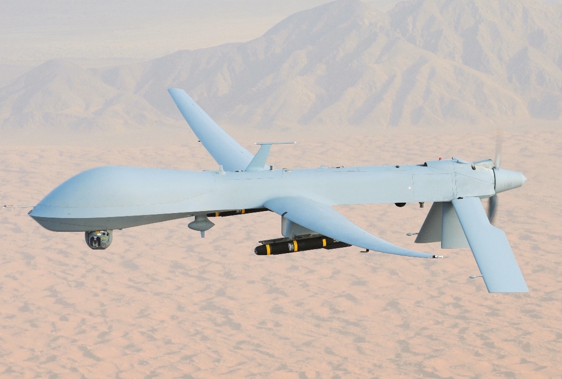A light grey-coloured drone carrying black missiles flies over a desert landscape.
