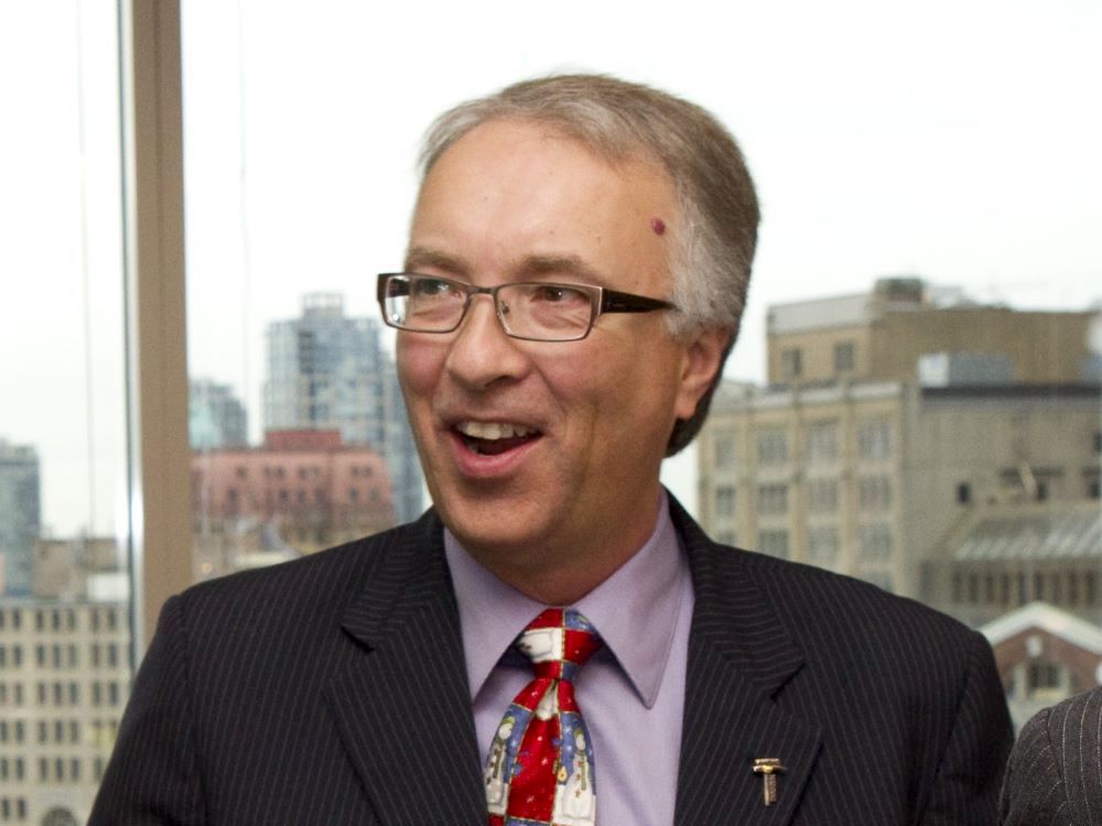 A man in late middle age with light skin tone and thinning grey and brown hair wears a suit with a purple shirt and colourful tie.