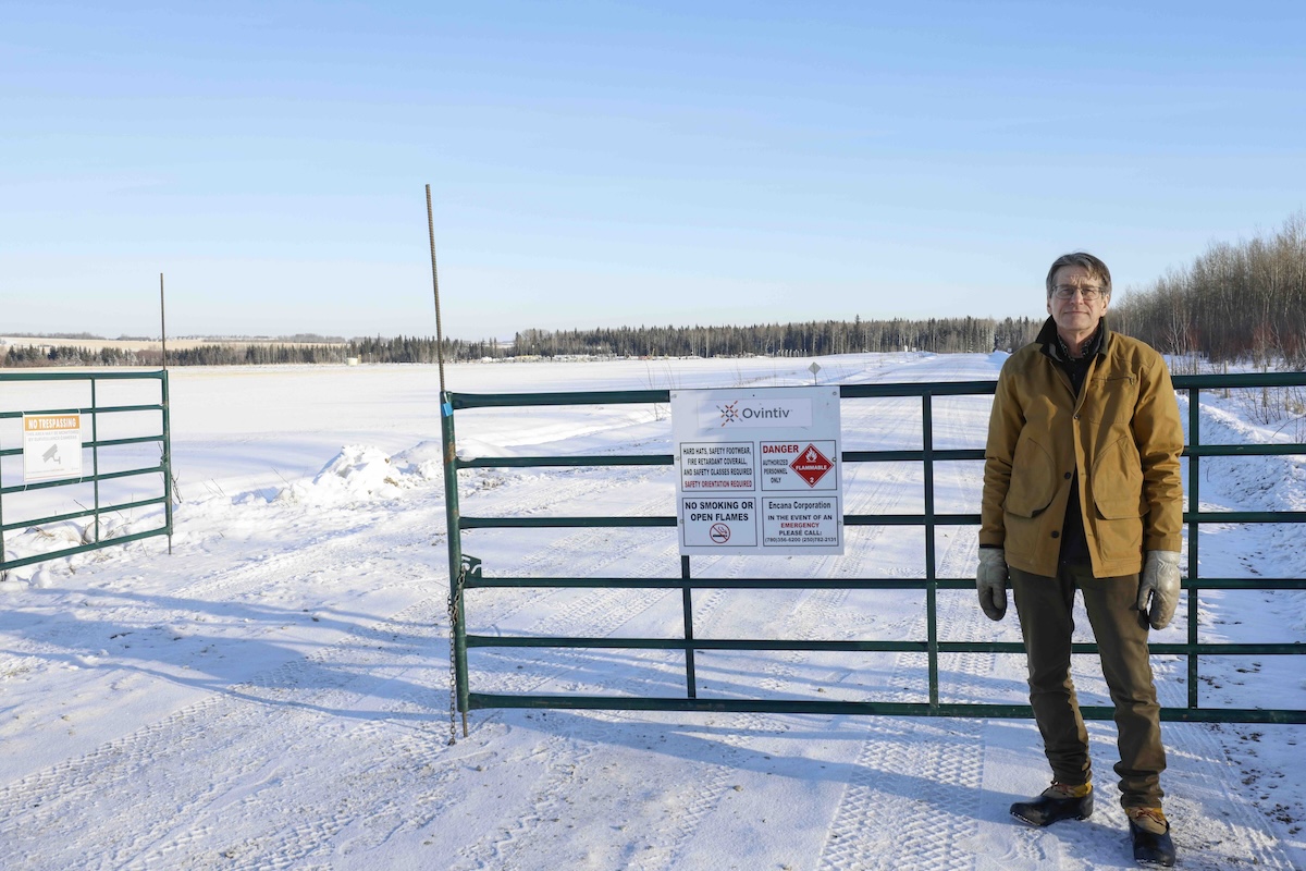 A light-skinned man in winter clothes stands beside a gate on a snowy road.