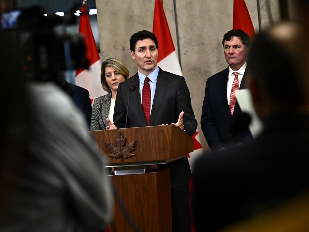 Justin Trudeau, a light-skinned man with short dark hair wearing a dark suit and red tie, speaks to media from a wooden podium with a maple leaf embossed on it.