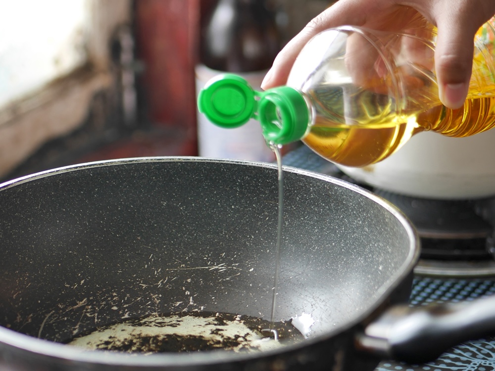 A person pours vegetable oil from a plastic bottle into a deep, scratched-up frying pan.