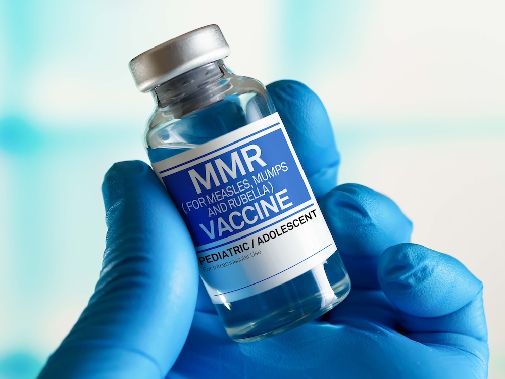 A hand in a blue latex glove holds a small glass vial with a silver cap. The blue and white label reads 'MMR (for measles, mumps and rubella) vaccine.' The background is out of focus and light blue.