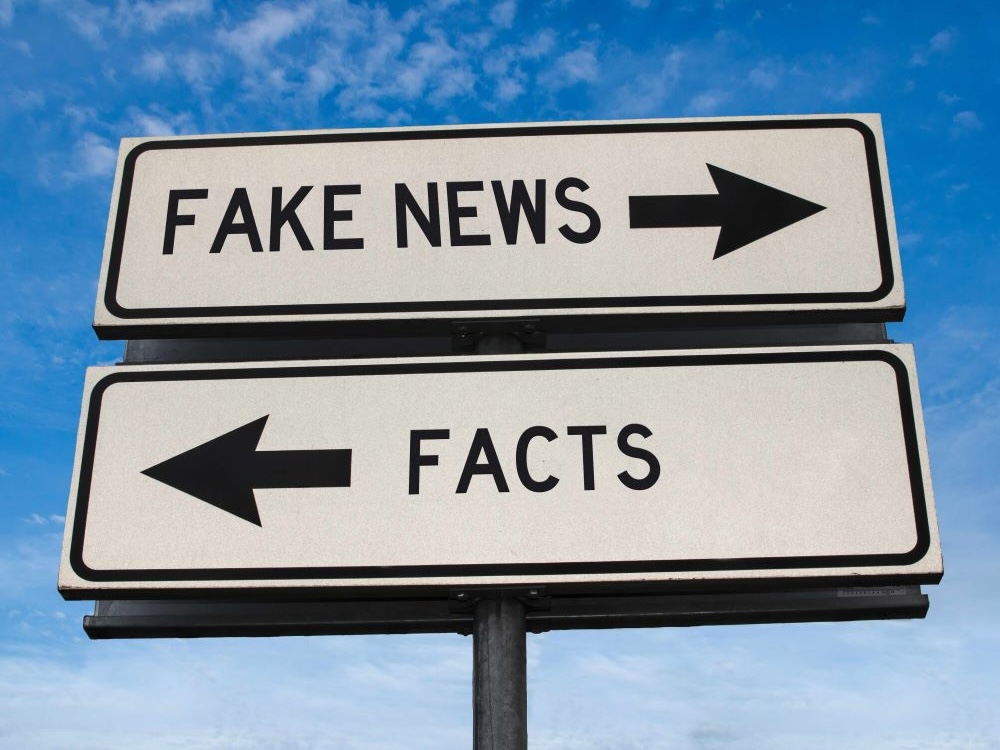 A signpost stands against a blue sky, shot from below. Two white signs with black text are attached to the post. The top sign reads “Fake News” and points right with an arrow; the bottom sign reads “Facts” and points left with an arrow.