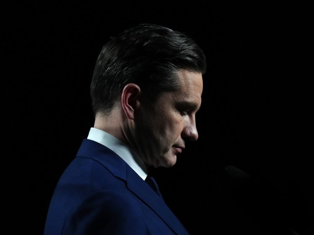 Against a black background, a profile view of Pierre Poilievre, a man with light skin tone and short dark hair wearing a dark blue suit and tie. He is looking downward with his eyes closed.