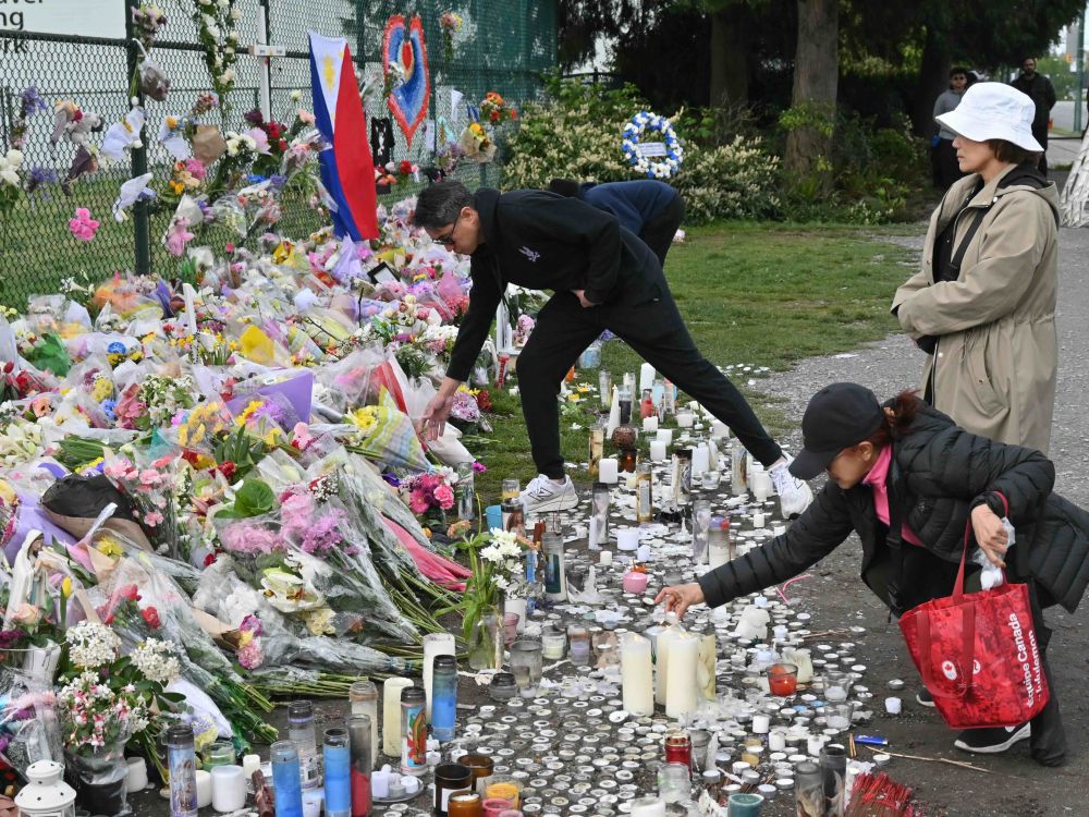Two people add bouquets to a large memorial, while a third looks on. There is a long row of flowers and candles.