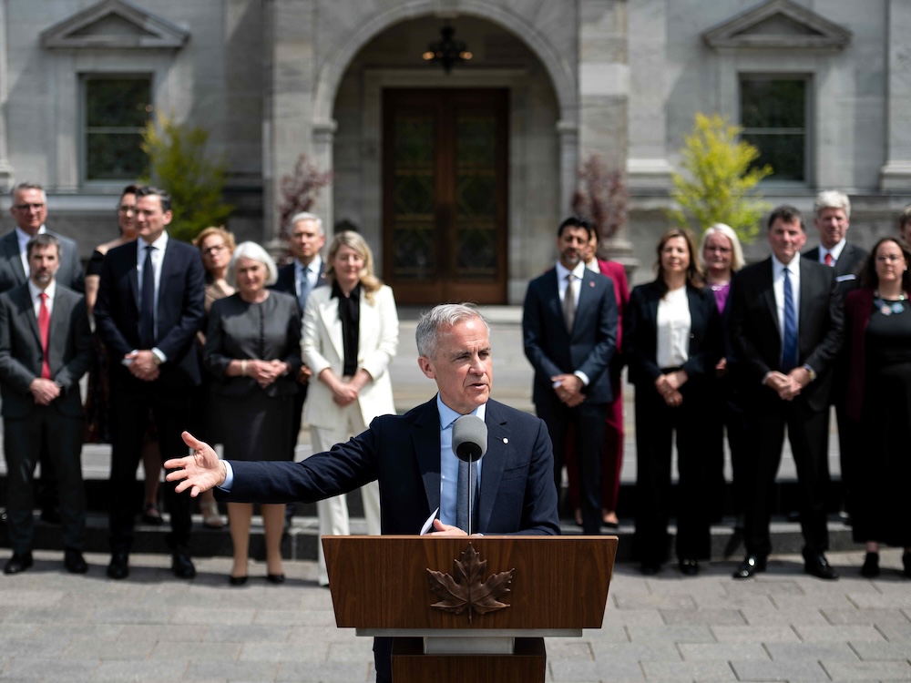 Mark Carney, a 60-ish light-skinned man, stands at a lectern, with his cabinet behind him.