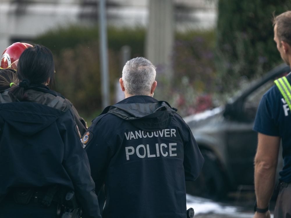 Two police officers and an emergency service worker stand with their backs to the camera. The police officer in the centre of the photo has short grey hair and is wearing a hooded jacket that reads ‘Vancouver Police.’ In front of them is the outline of a car razed by fire.
