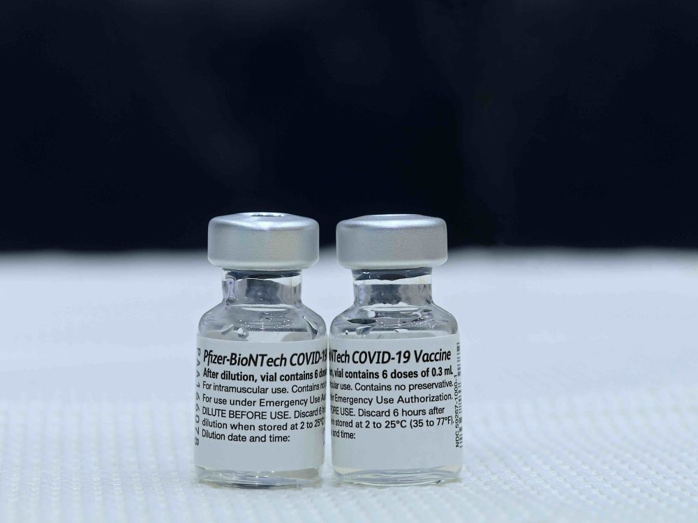 Two short, clear glass vials with silver lids sit next to each other on white fabric against a black background. Their black and white labels show they contain the Pfizer-BioNTech COVID-19 vaccine.
