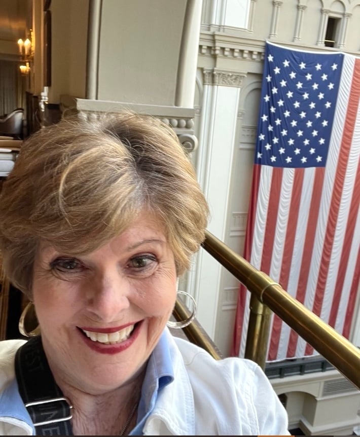 A 50-ish light-skinned woman with blondish hair smiles at the camera. A large U.S. flag is hanging vertically behind her.