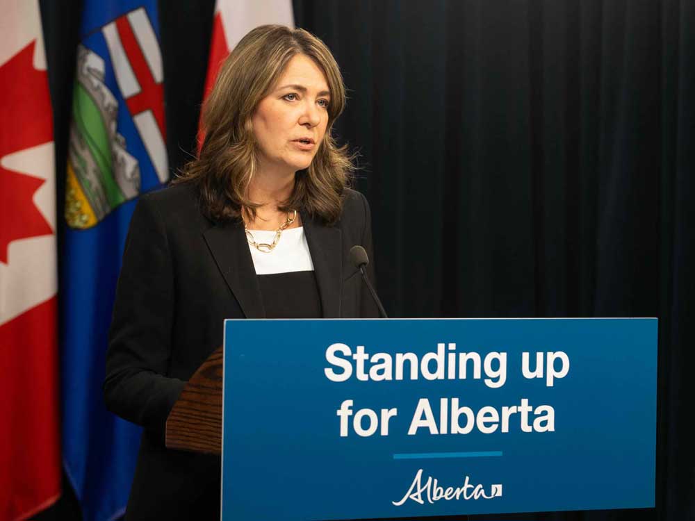 Danielle Smith has wavy, shoulder-length brown hair and a light skin tone. She is wearing a black blazer over a white top and standing behind a lectern on which a blue sign is affixed that reads “Standing up for Alberta.” Behind her are two Canada flags flanking one Alberta flag.