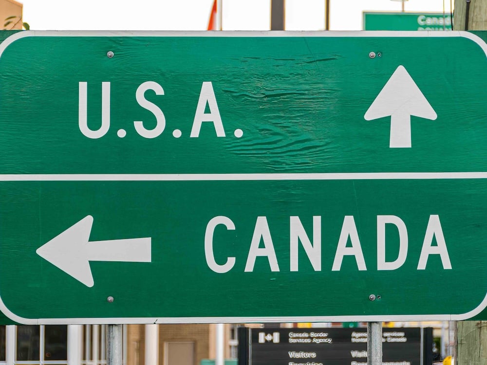 A green highway sign with white text reads ‘U.S.A.’ with an arrow pointing up, and ‘Canada’ with an arrow pointing to the left.