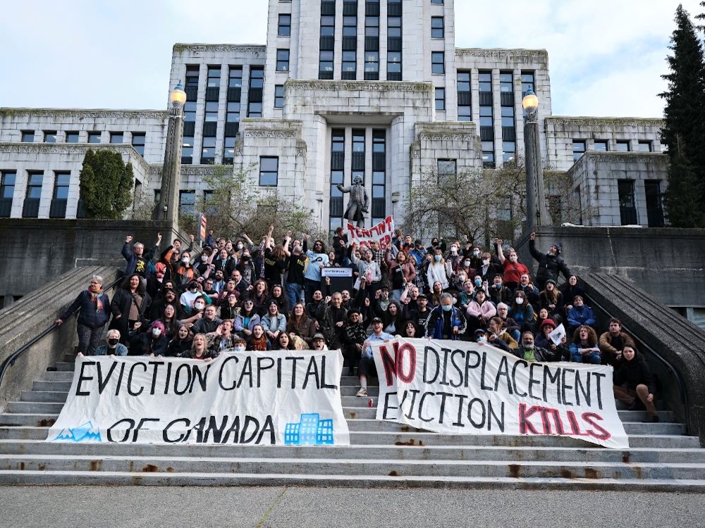 A group of people sit on the concrete steps in front Vancouver City Hall with large banners reading ‘Eviction Capital of Canada’ and ‘No Displacement. Eviction Kills.’