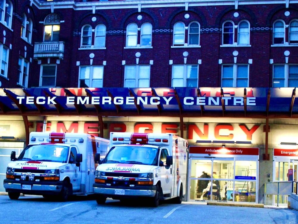 Two ambulances are parked under an awning that reads “Teck Emergency Centre” in capital letters. Through clear doors that say “Ambulance Entry Only,” people are seated and standing in a waiting room.