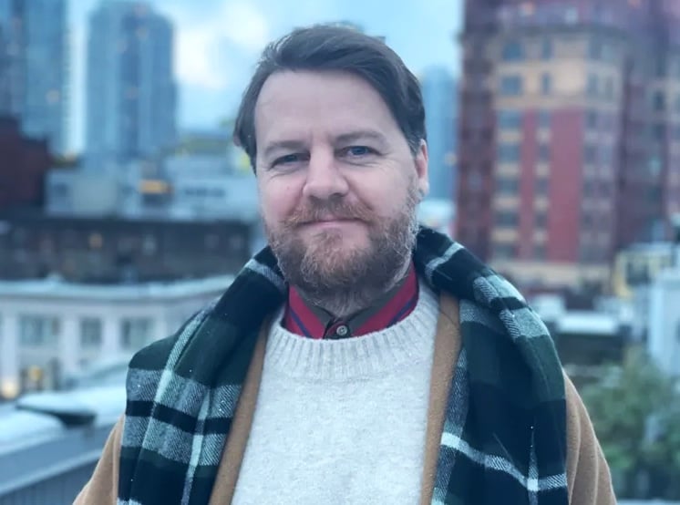 A 47-year-old light-skinned man with brown hair and a trimmed beard looks at the camera. He is wearing a jacket, a white sweater and a scarf draped over his shoulders.