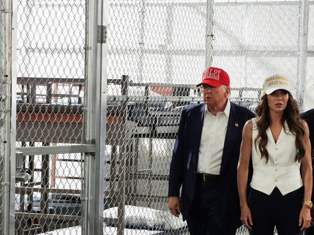 Donald Trump, in a red Gulf of America hat and wearing a blue suit with no tie, walks beside Kristi Noem, in a white MAGA hat. They are in front of cages used to hold detainees.