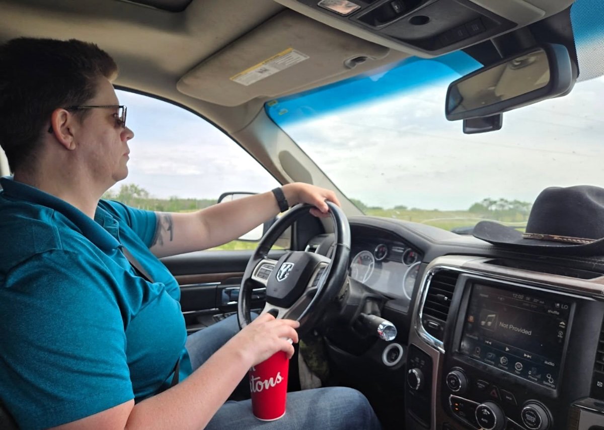 Bonnie Critchley, a light-skinned woman with short brown hair wearing jeans and a turquoise polo shirt, is seen viewed from side as she drives a car. She is holding a red Tim Hortons cup, and a black cowboy hat sits on the dashboard.