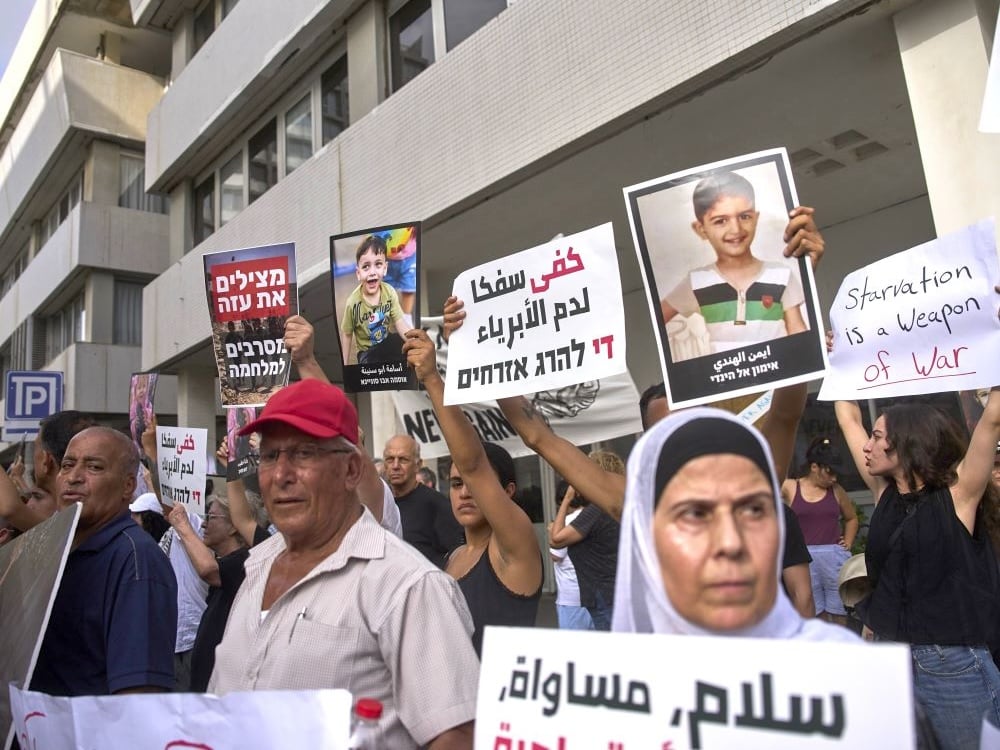 Protesters hold up signs of children and messages. One reads, ‘Starvation is a weapon of war.’