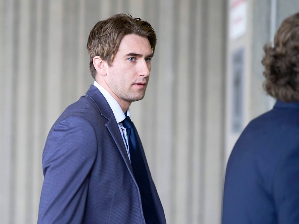 Michael McLeod is a man in his 20s with brown hair, a pale skin tone, blue eyes and a navy blue suit. He is walking with another person in a suit against a grey background.