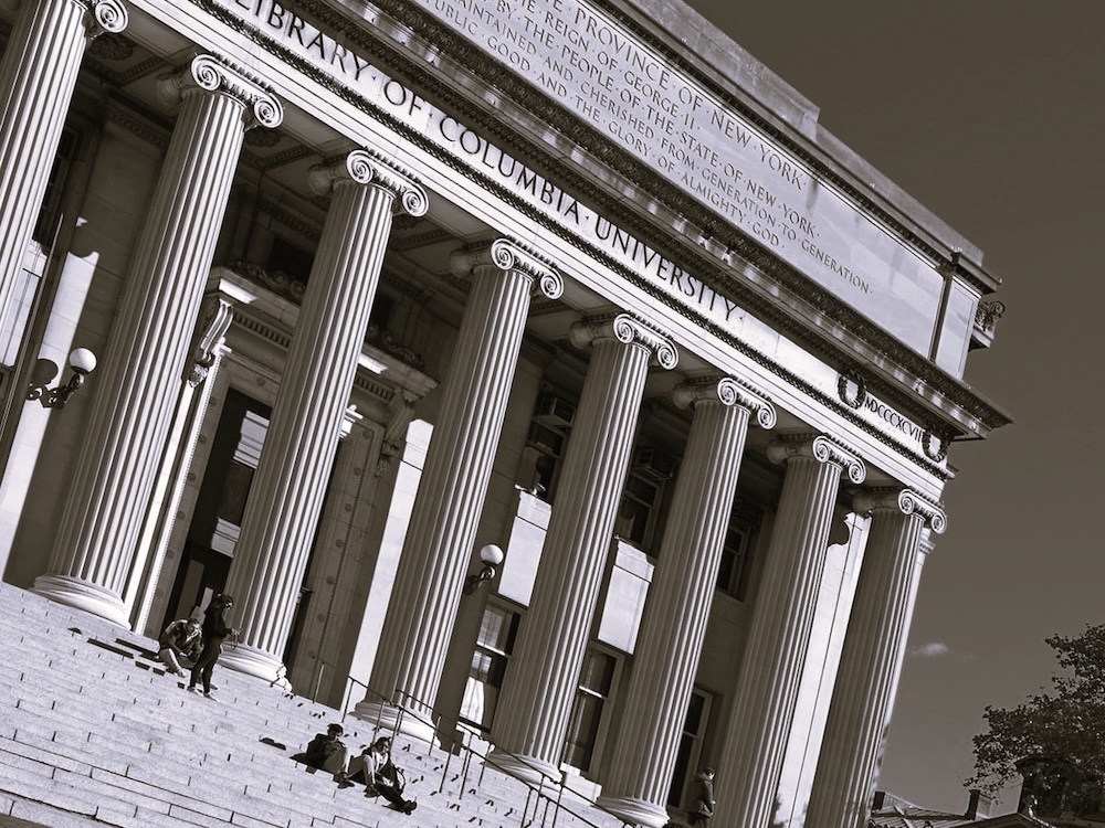 The Columbia University library is a wide beige building buttressed by Ionic columns. Two small groups of people are sitting on the front steps.