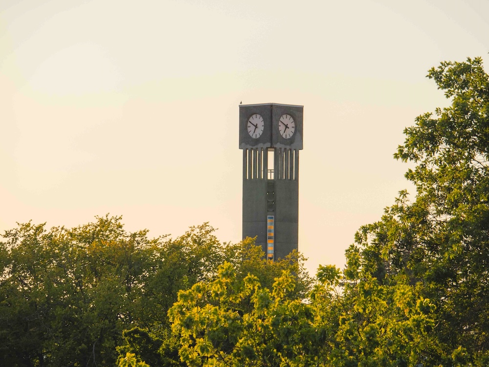 A tall concrete clock tower emerges above a stand of deciduous trees against a yellow, hazy sky.