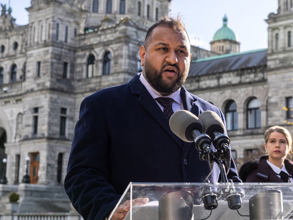 Ravi Parmar stands at a lectern in front of the provincial legislature in Victoria. He has medium-dark skin tone, a dark beard, and short dark hair. 